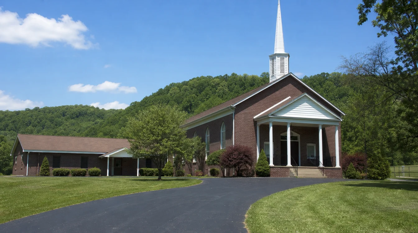 An unnamed small West Virginia church of red-brown brick with a modest white entry portico and a connected low fellowship annex, leafy Appalachian foothills in the distance and a normal paved approach